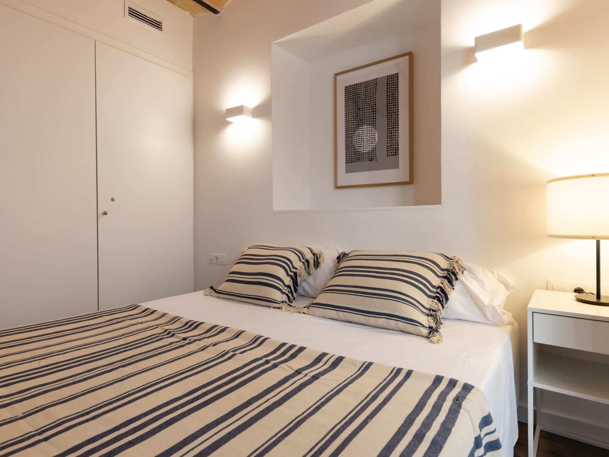 A bedroom with a bed, two pillows, a bedside table, a lamp, a framed artwork, and a wardrobe.