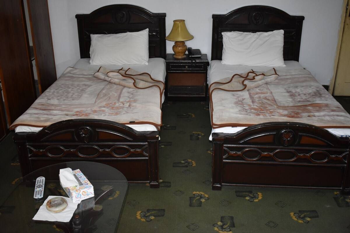 A hotel room with two wooden beds, each with a white pillow and a patterned bedspread. There is a small table with a lamp and a remote control.