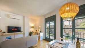 Ein modernes Wohnzimmer mit Flachbildfernseher, einer Couch, einem Esstisch für zwei Personen und großen Fenstern mit Blick ins Freie.