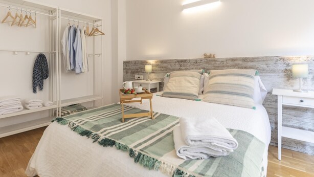 Ein ordentlich eingerichtetes Schlafzimmer mit Bett, Nachttischen, einer Kommode und einem Kleiderschrank sowie einer Kleiderstange mit Kleidung.
