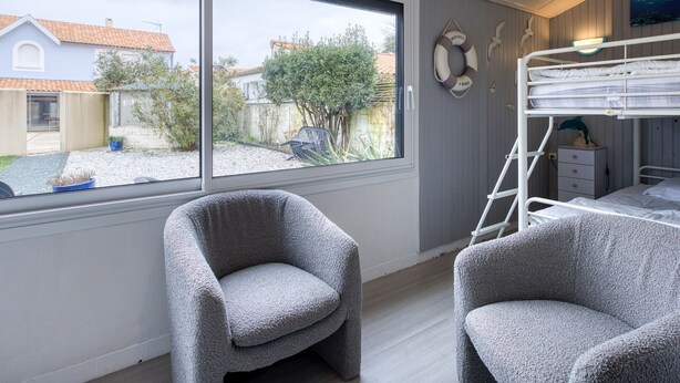 Una habitación con una cama nido, dos sillones grises, un ventanal con vista a un jardín y casas, y un salvavidas colgado en la pared.