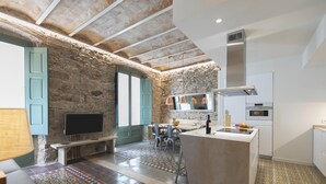 A spacious living area with a stone wall, a TV, a dining table, and a kitchen with modern appliances. - Bravissimo Mercaders 3-1-2, unique and with 3 bedrooms in the historic center (Girona)