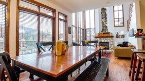 A spacious living area with a large wooden table, comfortable seating, a stone fireplace, and large windows offering a view. - Spacious multi level 4 bedroom + den townhouse, ski in/ski out (Sun Peaks)