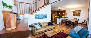 A modern living room with a sofa, coffee table, and a dining area with chairs. A staircase leads to an upper level.