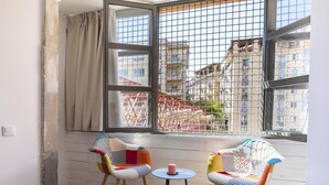 Ein Zimmer mit zwei Stühlen, einem kleinen Tisch, einem Fenster mit Blick auf Gebäude und einem Vorhang.