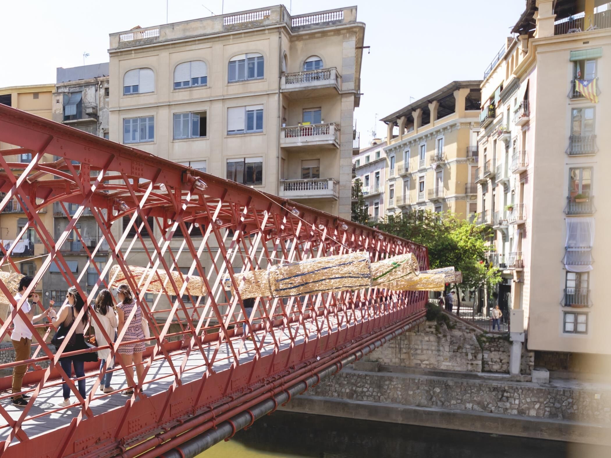 A red metal bridge with wooden planks across a stone riverbed, surrounded by multi-story buildings with balconies.