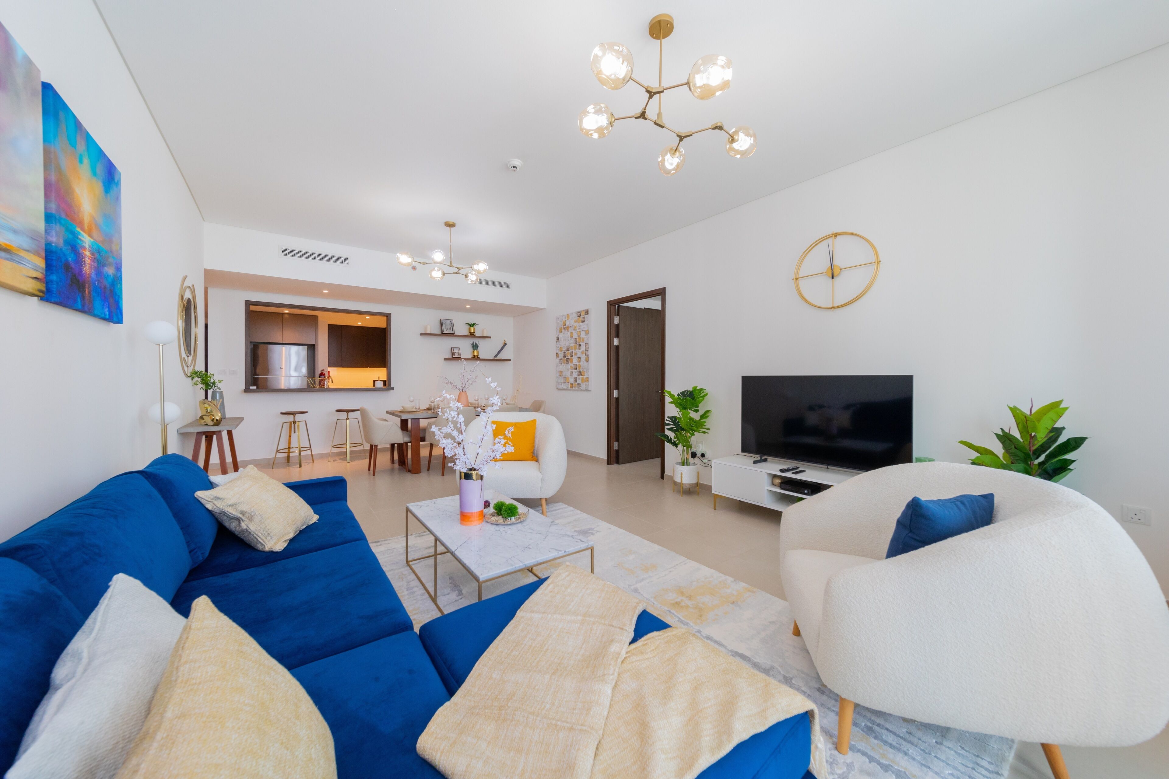 Een moderne woonkamer met een blauwe bank, een witte fauteuil en een televisie. Achtergrond is een keuken met een eethoek.