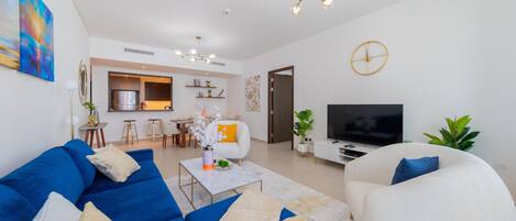 Een moderne woonkamer met een blauwe bank, een witte fauteuil en een televisie. Achtergrond is een keuken met een eethoek.