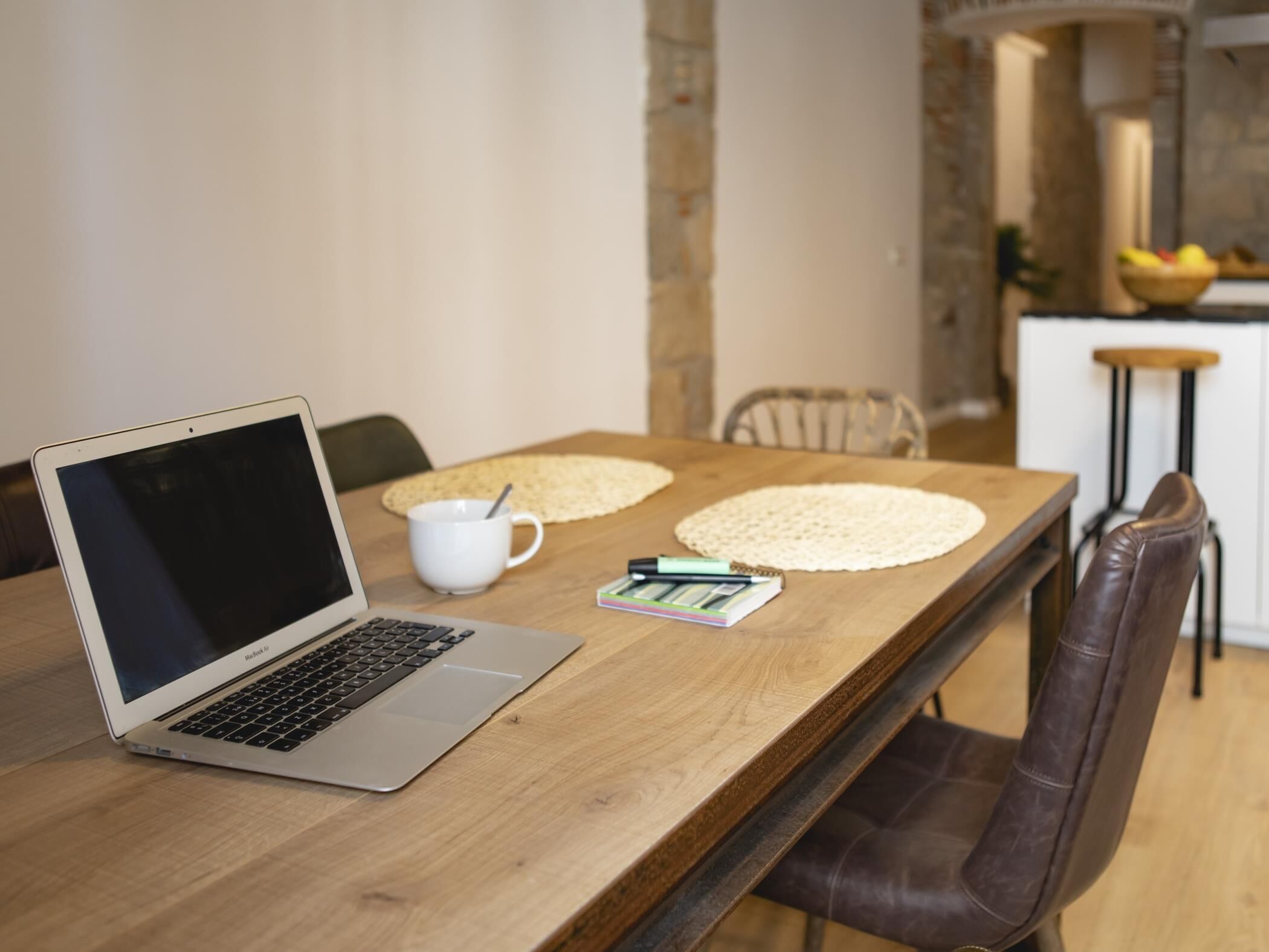 A wooden dining table with a laptop, a white mug, and a notebook.