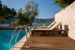 A wooden deck with a pool, lounge chairs, and a stone wall. - Liana's House with great view in Vlicho Bay & big private pool ! (Lefkada)