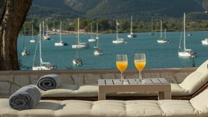 A seaside lounge with two glasses of orange juice on a table, a tree, and a view of boats on the water. - Liana's House with great view in Vlicho Bay & big private pool ! (Lefkada)
