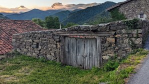 Un edificio rústico de piedra con puerta de madera, rodeado de abundante vegetación y un paisaje montañoso.