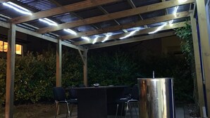 Een terras buiten met een metalen dak, een bargedeelte met stoelen en een afvalbak.
