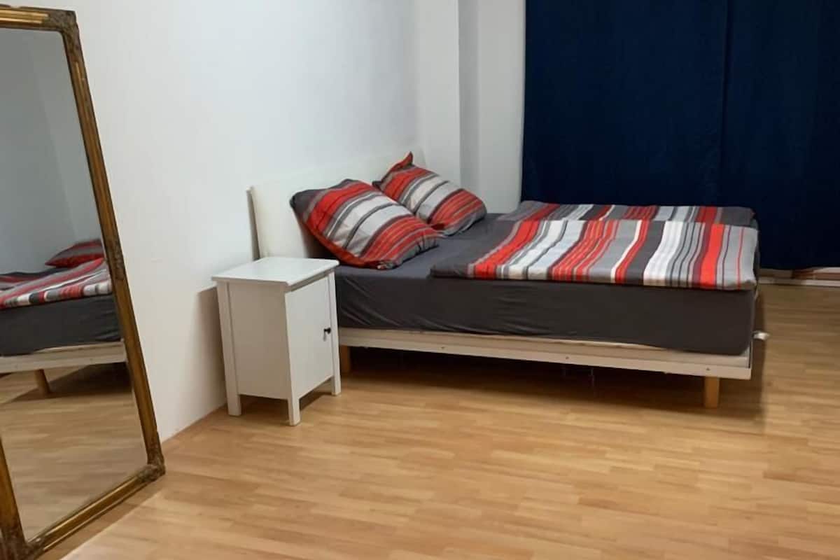 Een slaapkamer met houten vloer, een bed met gestreepte beddengoed, een witte nachtkastje en een spiegel.