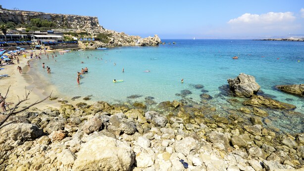 Ein felsiger Strand mit klarem türkisblauem Wasser, einige Badende und ein Schiff in der Ferne.