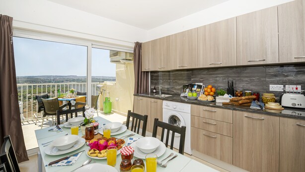 A modern kitchen with wooden cabinets, a built-in oven, and a dining area with a table set for breakfast. - Summer Breeze Superior Apartment with Terrace by Getaways Malta (Mellieha)
