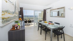 A dining area with a table set for four, a large painting on the wall, and a view of the city through the balcony doors. - Summer Breeze Penthouse with large Terrace and Hot Tub by Getaways Malta (Mellieha)