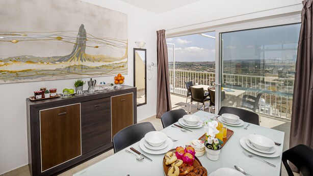 A dining area with a table set for four, a large abstract painting on the wall, and a view of the city through large windows. - Summer Breeze Penthouse with large Terrace and Hot Tub by Getaways Malta (Mellieha)