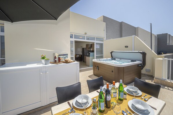 An outdoor dining area with a hot tub, a table set for four, and a kitchen area in the background.