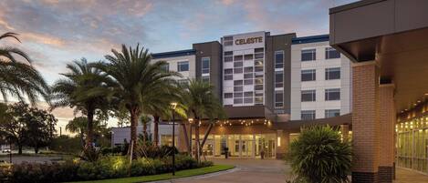A hotel with a modern design, featuring a prominent entrance, palm trees, and a well-lit facade.