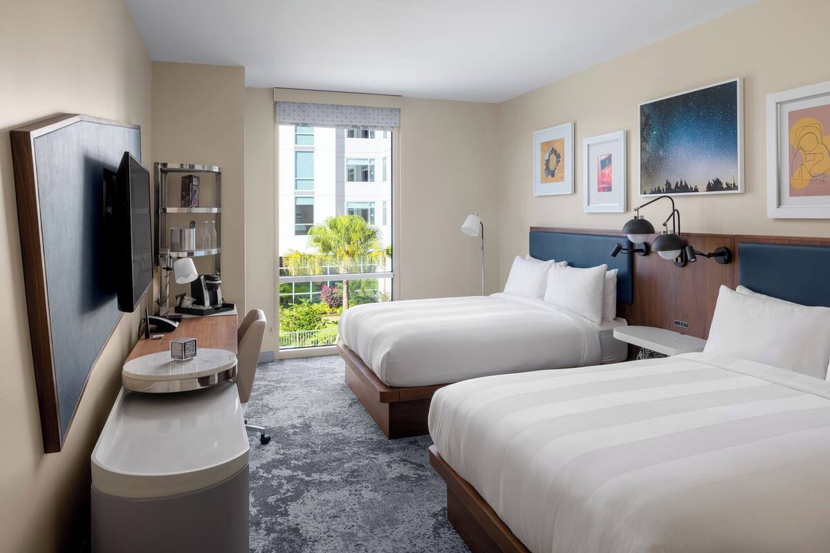 A hotel room with two beds, a desk with a chair, a TV, a bookshelf, and a balcony with a view of buildings and greenery.