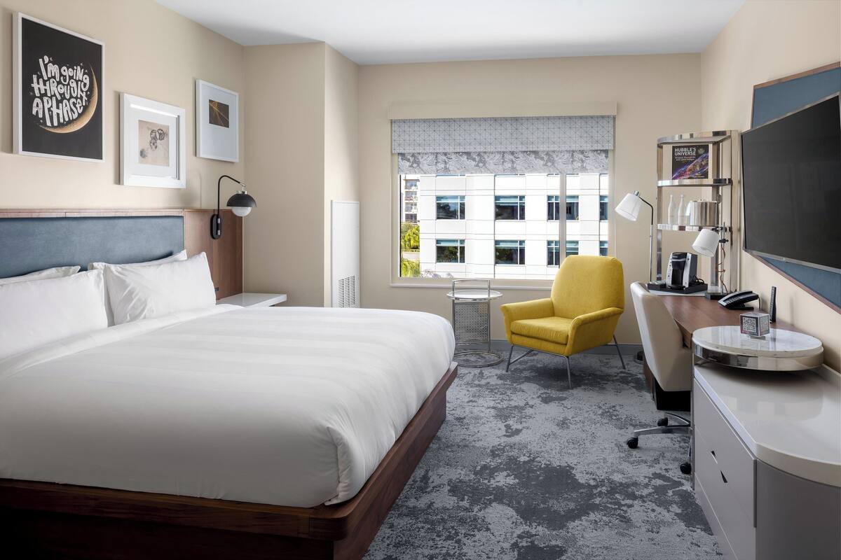 A hotel room with a large bed, a desk with a chair, a TV, and framed pictures on the wall.