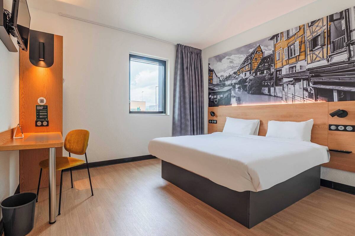 A hotel room with a bed, a desk, a chair, a window, and a mural on the wall.