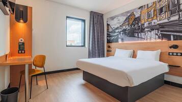 A hotel room with a bed, a desk, a chair, a window, and a mural on the wall.