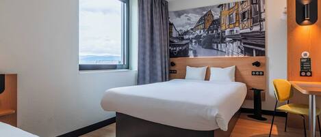 A hotel room with a bed, a desk, a chair, a window with curtains, a wall-mounted light, and a painting.