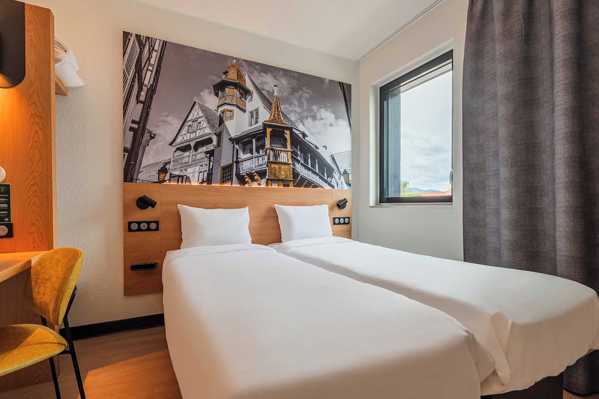 A hotel room with a bed, a desk, a chair, a painting of a building, and a window with curtains.