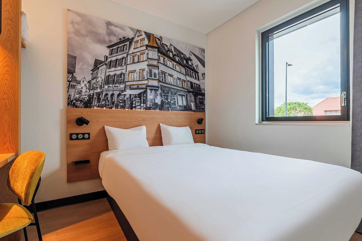 A hotel room with a bed, a headboard with controls, a window, and a wall-mounted artwork.