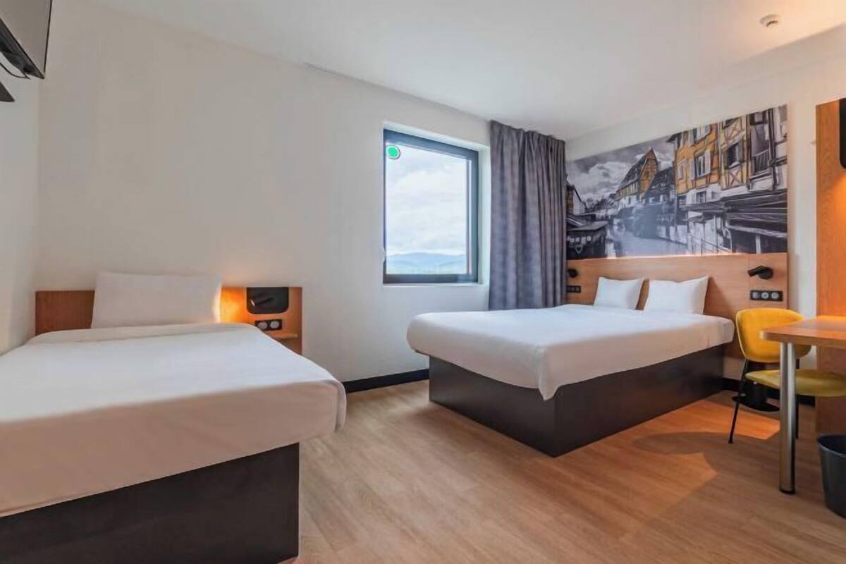 A hotel room with two beds, a window, a painting on the wall, and a small table with a chair.
