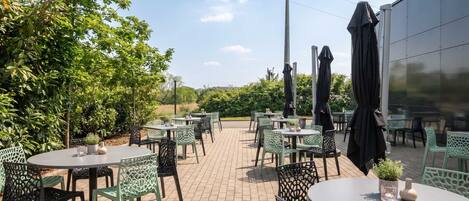 Ein Terrassenbereich im Freien mit runden Tischen und Stühlen, umgeben von viel Grün und blauem Himmel.