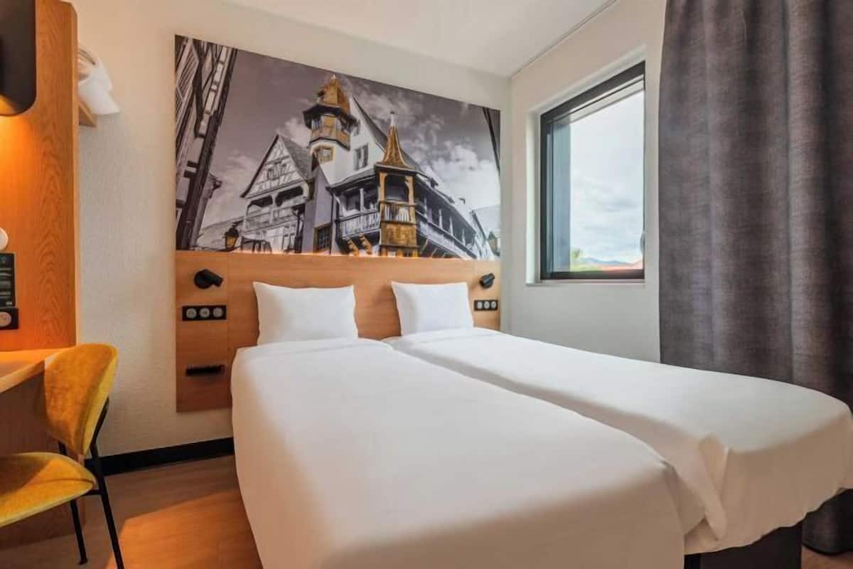 A hotel room with a bed, a desk, a chair, a large window, and a painting on the wall.