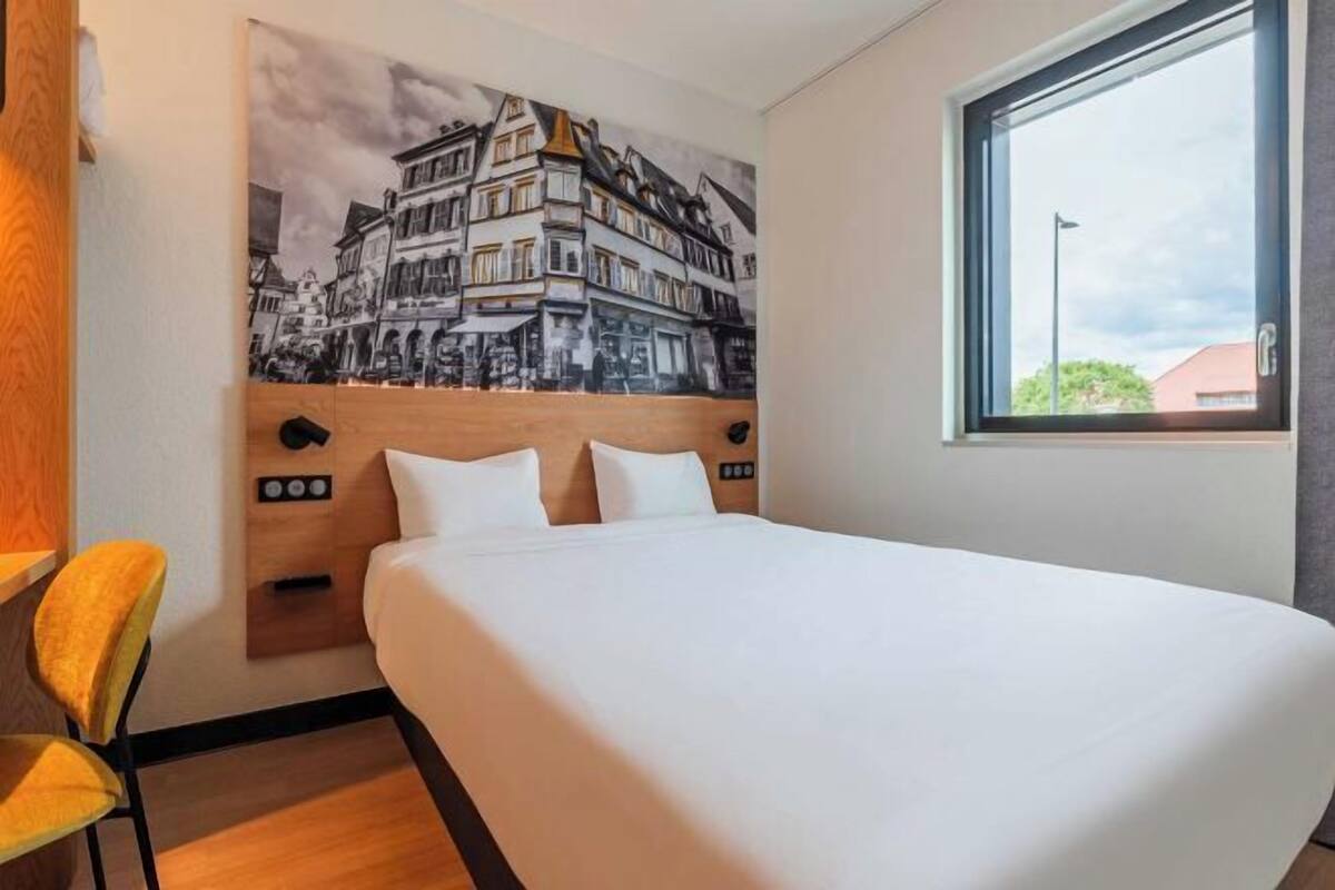 A hotel room with a bed, a chair, a window, and a wall-mounted artwork.