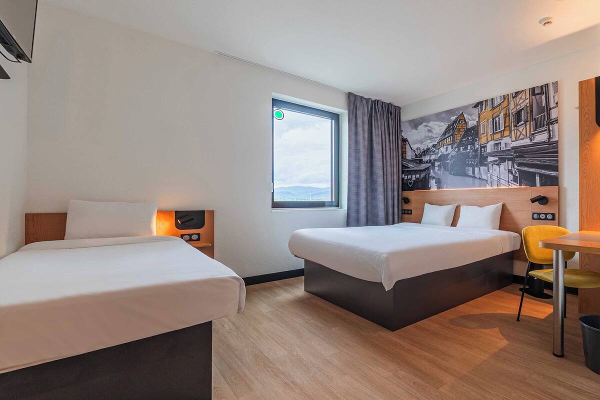 A hotel room with two beds, a window with curtains, a painting on the wall, a small table, and a chair.