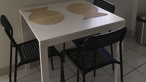 Una mesa blanca con dos individuales tejidos, sillas negras y una ventana con celosía metálica.