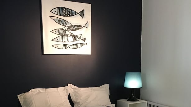 Una cama bien tendida con ropa de cama a rayas, un cuadro de peces colgado en la pared, una lámpara de noche y una mesita de noche blanca.