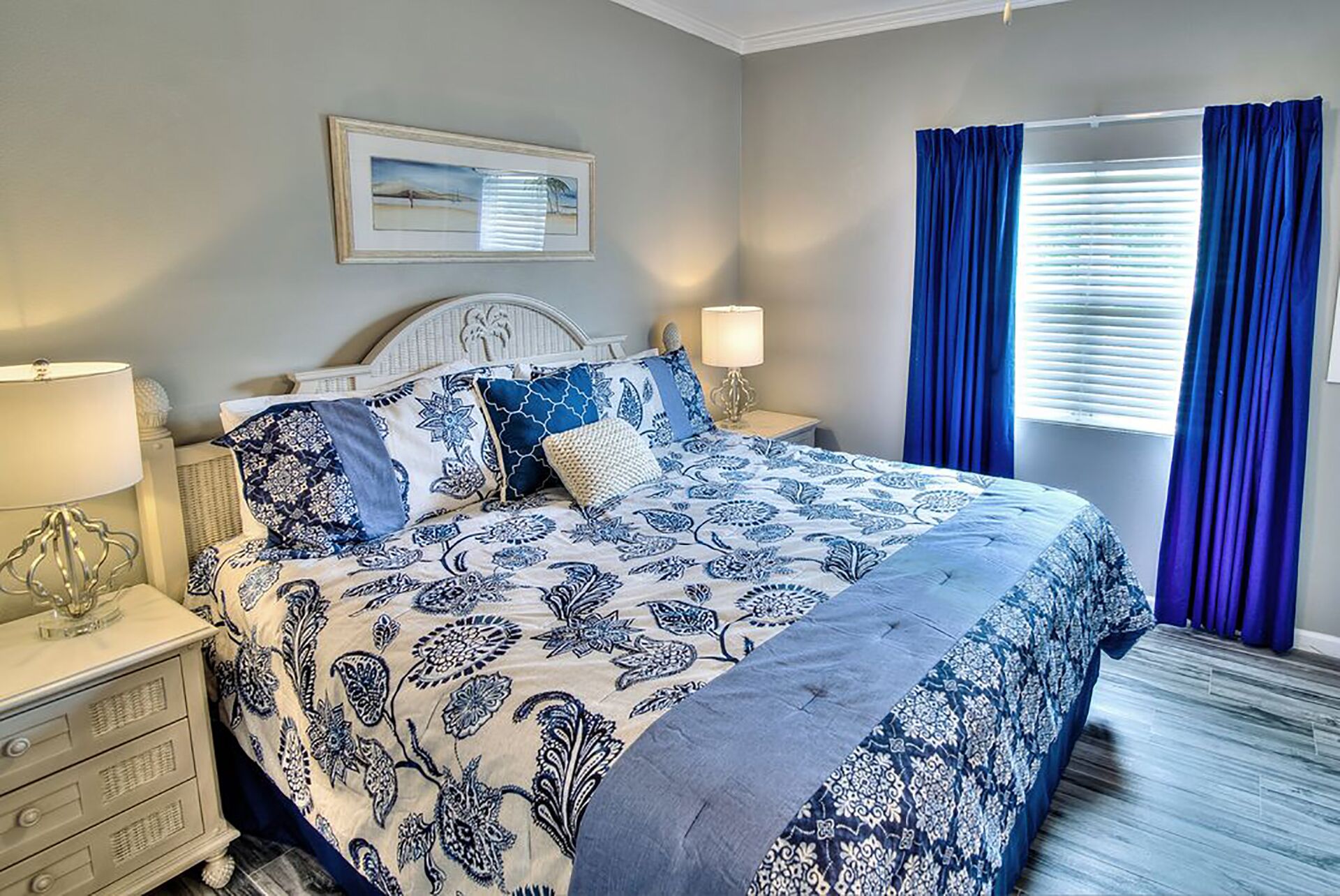 A bedroom with a bed, nightstands, a lamp, a picture on the wall, and blue curtains.