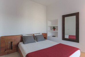 A bedroom with a wooden headboard, a large bed with white bedding and a red throw, a small shelf with a plant, and a large mirror with a geometric frame.