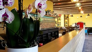 A bar area with a white orchid plant, wooden ceiling, hanging pendant lights, and shelves with bottles and glasses. - Ludwig Hotel (Martonvásár)