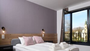 A modern hotel room with a large bed, wooden headboard, bedside lamps, and a view of a building through a large window. - Ludwig Hotel (Martonvásár)