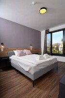 Quarto de hotel moderno com uma cama grande, cabeceira de madeira, abajures e vista para um edifício por uma janela ampla.