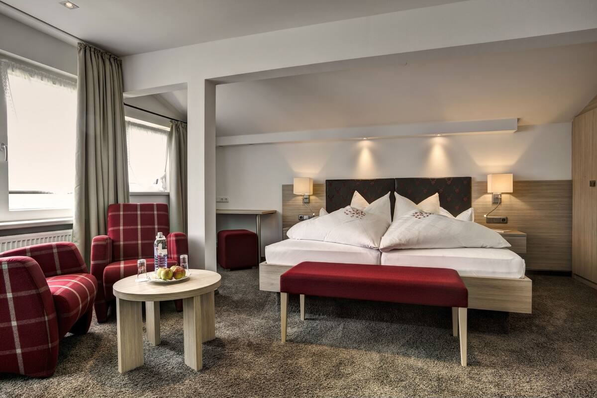 A hotel room with a bed, two red armchairs, a small round table with a bottle and glasses, and a red bench.