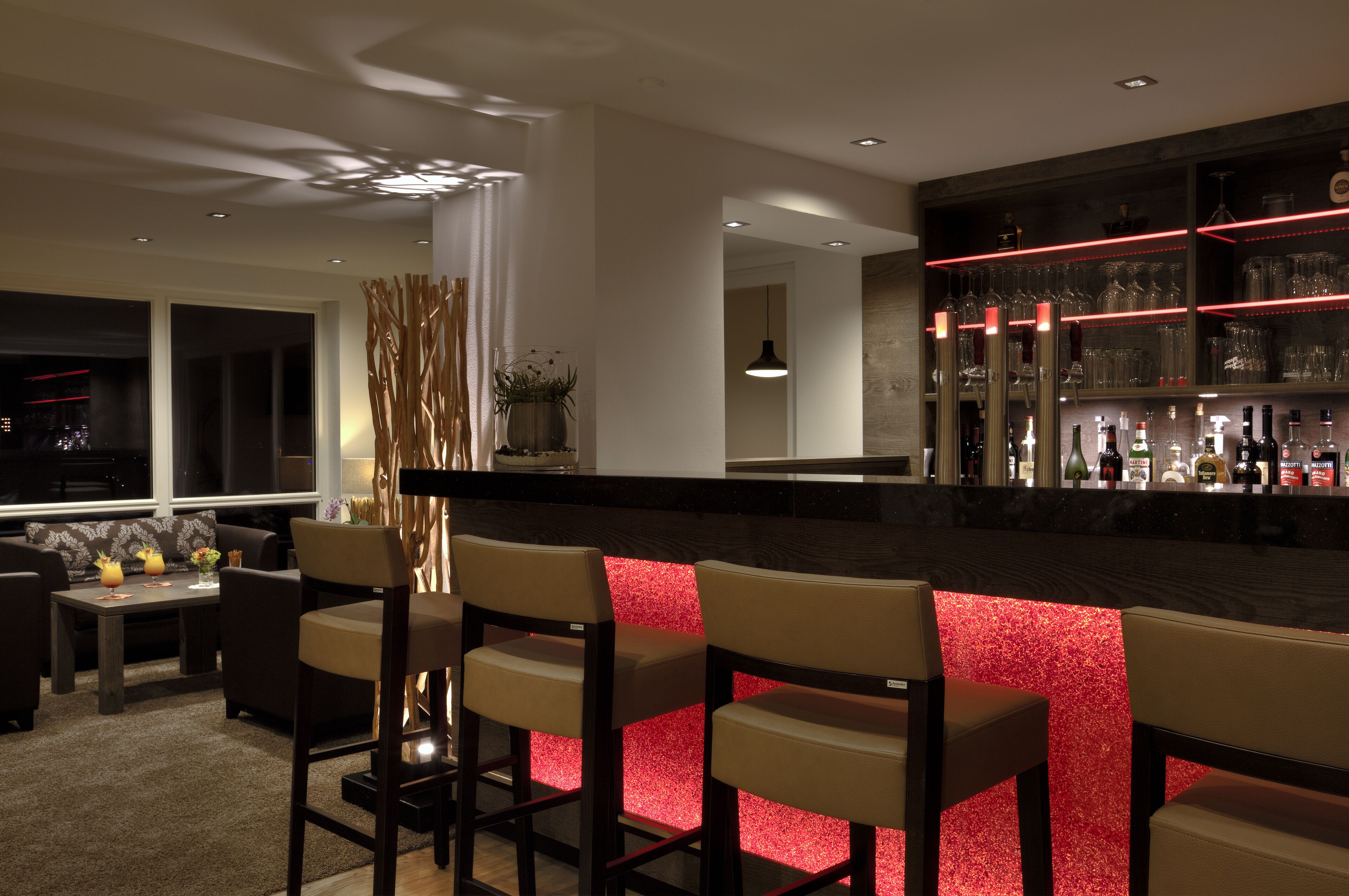 A modern bar area with a red-lit bar counter, seating, and a view of the city at night.