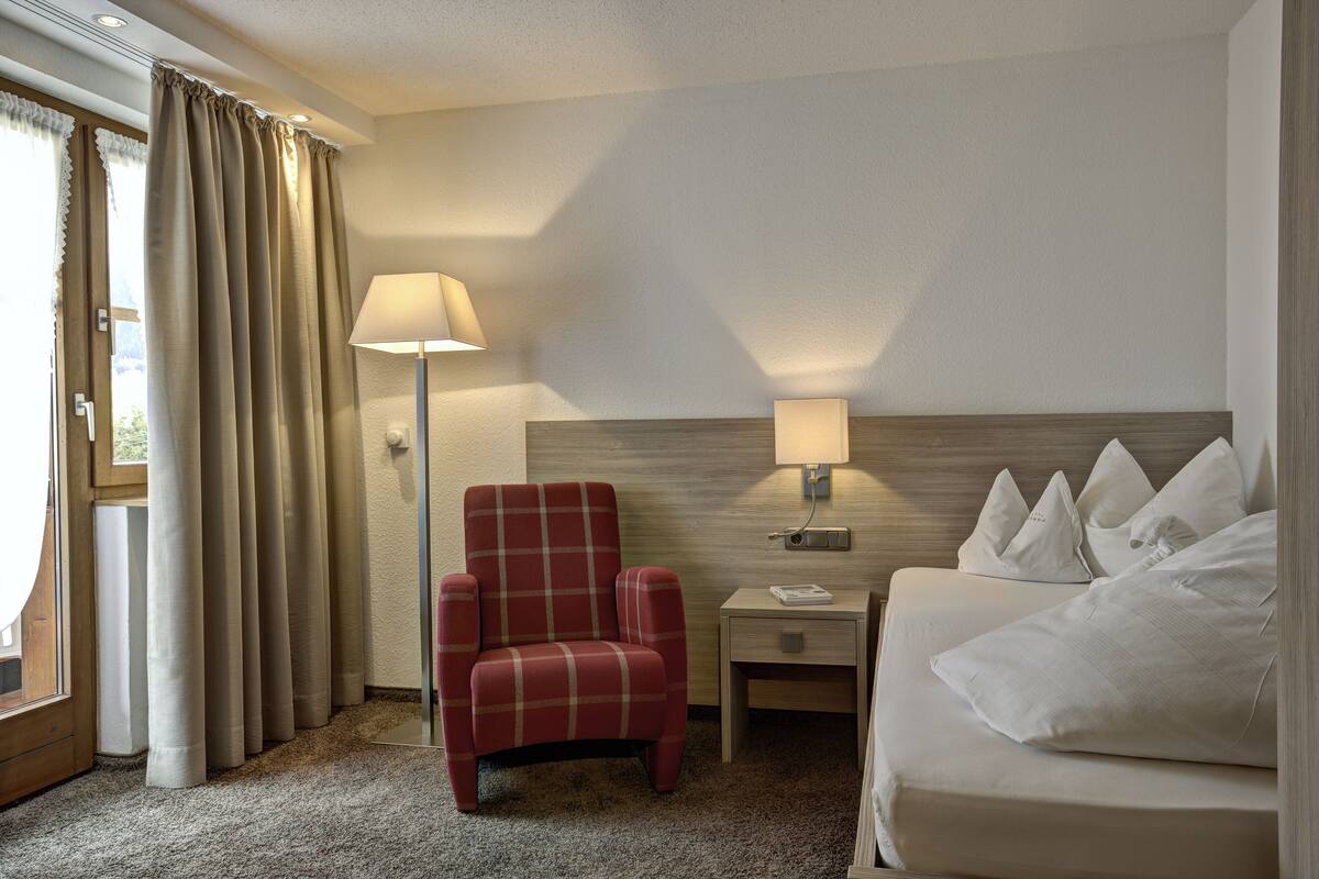 A hotel room with a bed, a chair, a nightstand, a lamp, and a window with curtains.