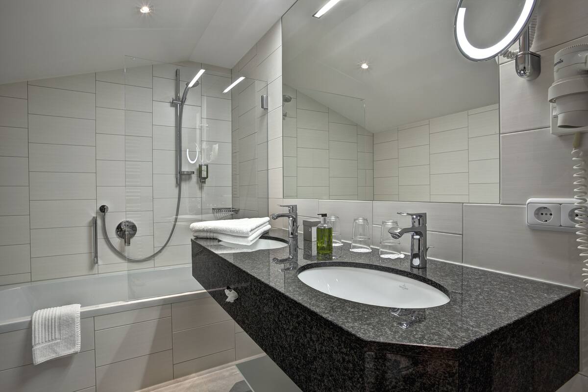 A modern bathroom with a large mirror, a bathtub, a sink with a granite countertop, and a handheld showerhead.