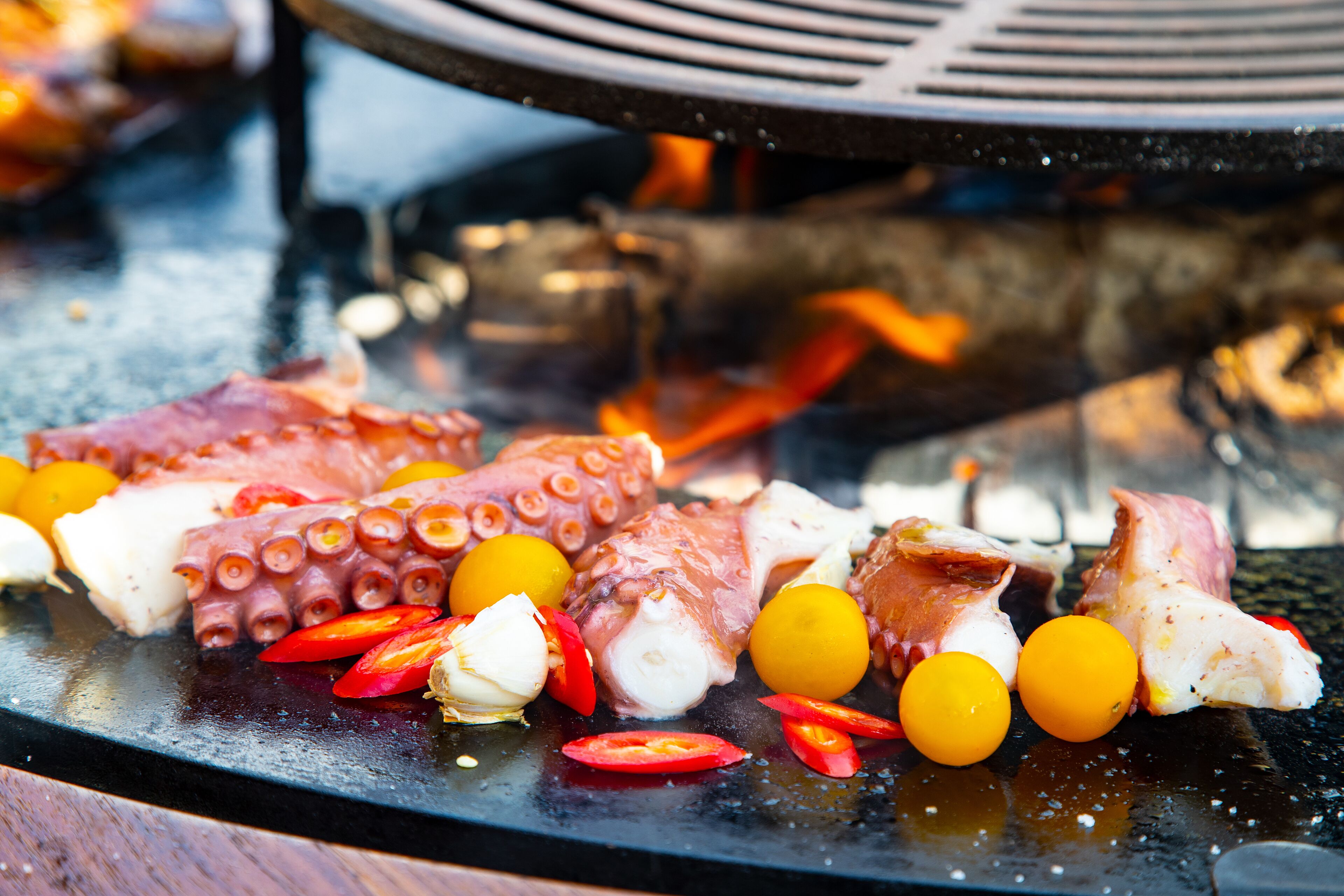 A grill with octopus tentacles, garlic cloves, and cherry tomatoes being cooked.