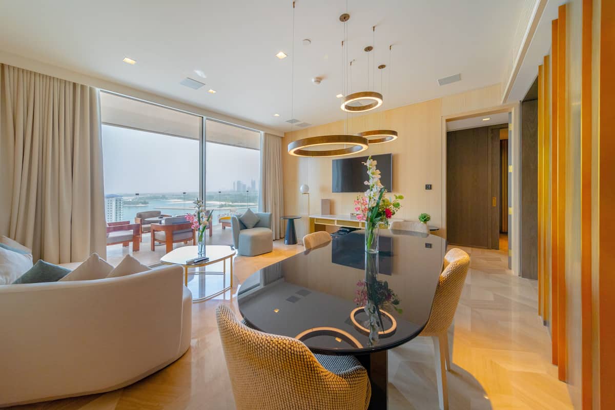 Una habitación de hotel moderna con un ventanal grande, un comedor con mesa de vidrio y una sala de estar con sofá y sillones.