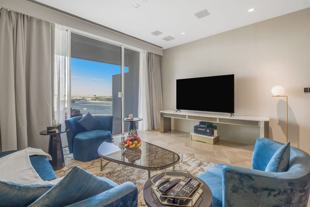 Una sala moderna con un televisor de pantalla plana grande, asientos cómodos, una mesa de centro con controles remotos y vistas a la ciudad a través de ventanas amplias.